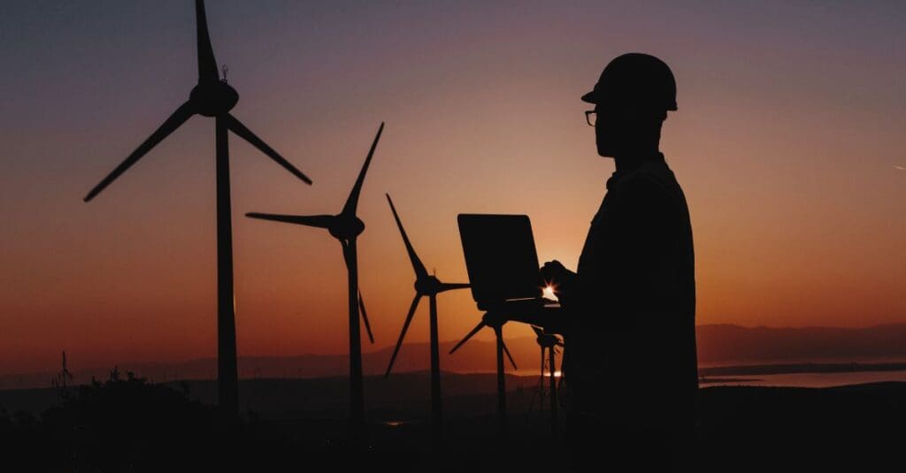 A professional working on a laptop in front of wind turbines during sunset, symbolising the intersection of technology and renewable energy. Navitas Resourcing Group connects top talent to clean energy projects like wind energy.