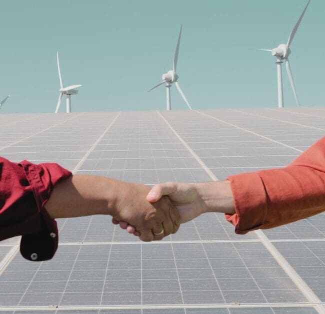 Two clean energy professionals shaking hands in front of solar panels and wind turbines, symbolising collaboration in the renewable energy sector. Navitas Resourcing Group connects top talent with projects that drive the future of sustainable energy.