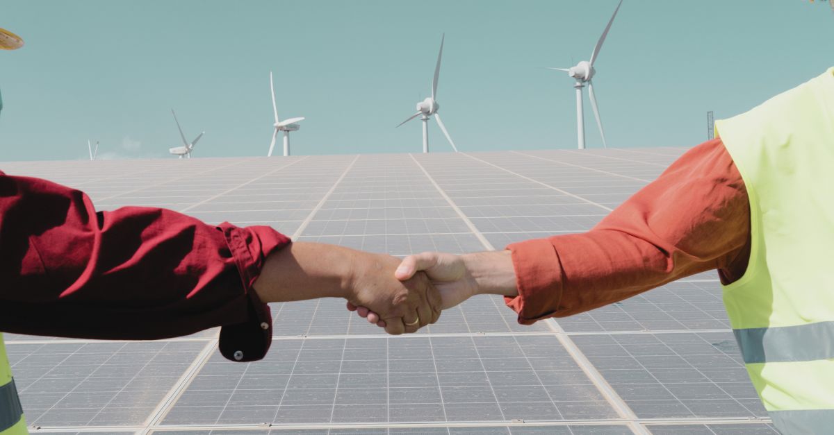 Two clean energy professionals shaking hands in front of solar panels and wind turbines, symbolising collaboration in the renewable energy sector. Navitas Resourcing Group connects top talent with projects that drive the future of sustainable energy.