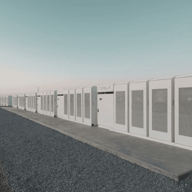 A large-scale Tesla battery storage facility with a wind turbine in the background, highlighting the role of energy storage in stabilising renewable power. Navitas Resourcing Group connects skilled professionals to the energy storage sector, supporting the clean energy transition.