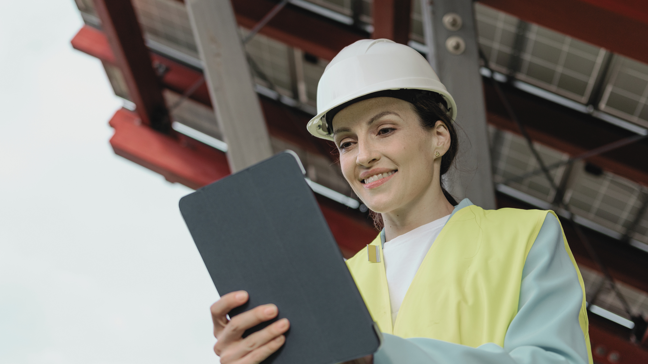 Female engineer wearing a hard hat and high-vis vest, reviewing a digital tablet beneath solar panels - symbolising the growing demand for skilled talent in the clean energy sector.