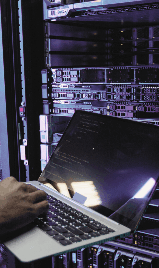 An engineer working on a laptop in a data centre server room, maintaining energy and infrastructure systems.