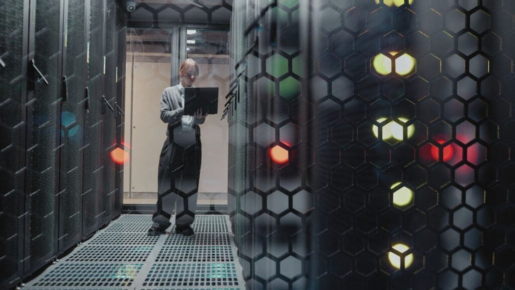 Engineer using a laptop inside a data centre server room, surrounded by racks and digital infrastructure equipment