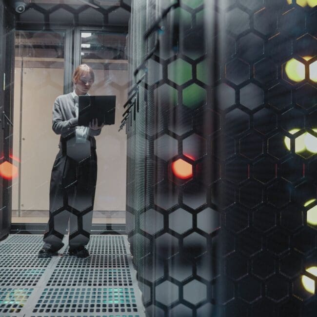 Engineer using a laptop inside a data centre server room, surrounded by racks and digital infrastructure equipment