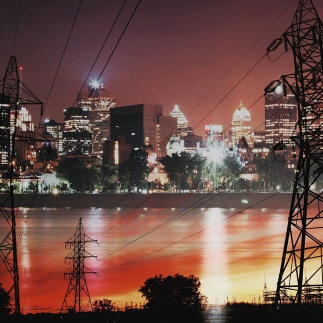Electricity transmission towers and power lines near a city skyline at night, representing grid infrastructure and energy supply