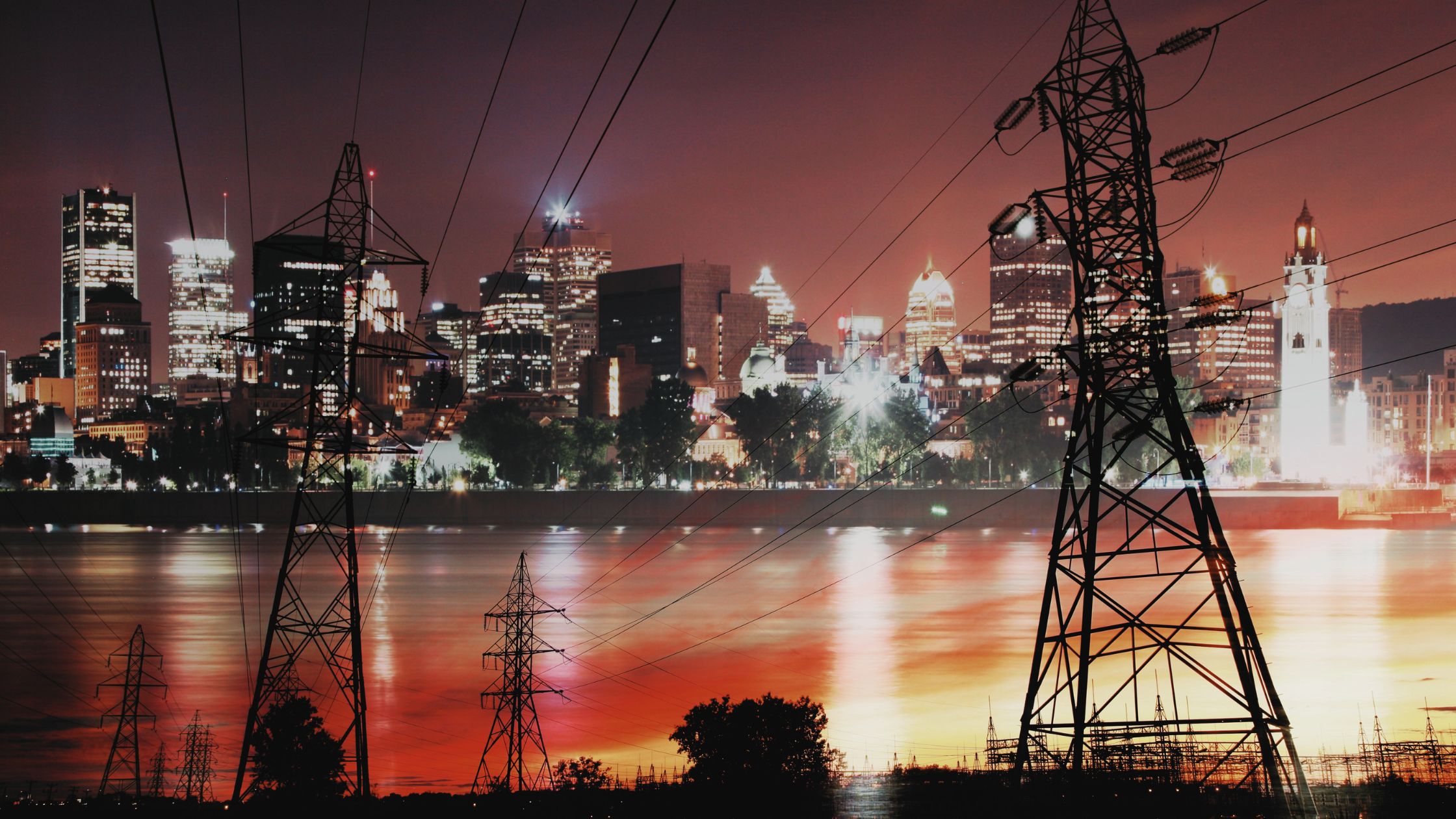 Electricity transmission towers and power lines near a city skyline at night, representing grid infrastructure and energy supply